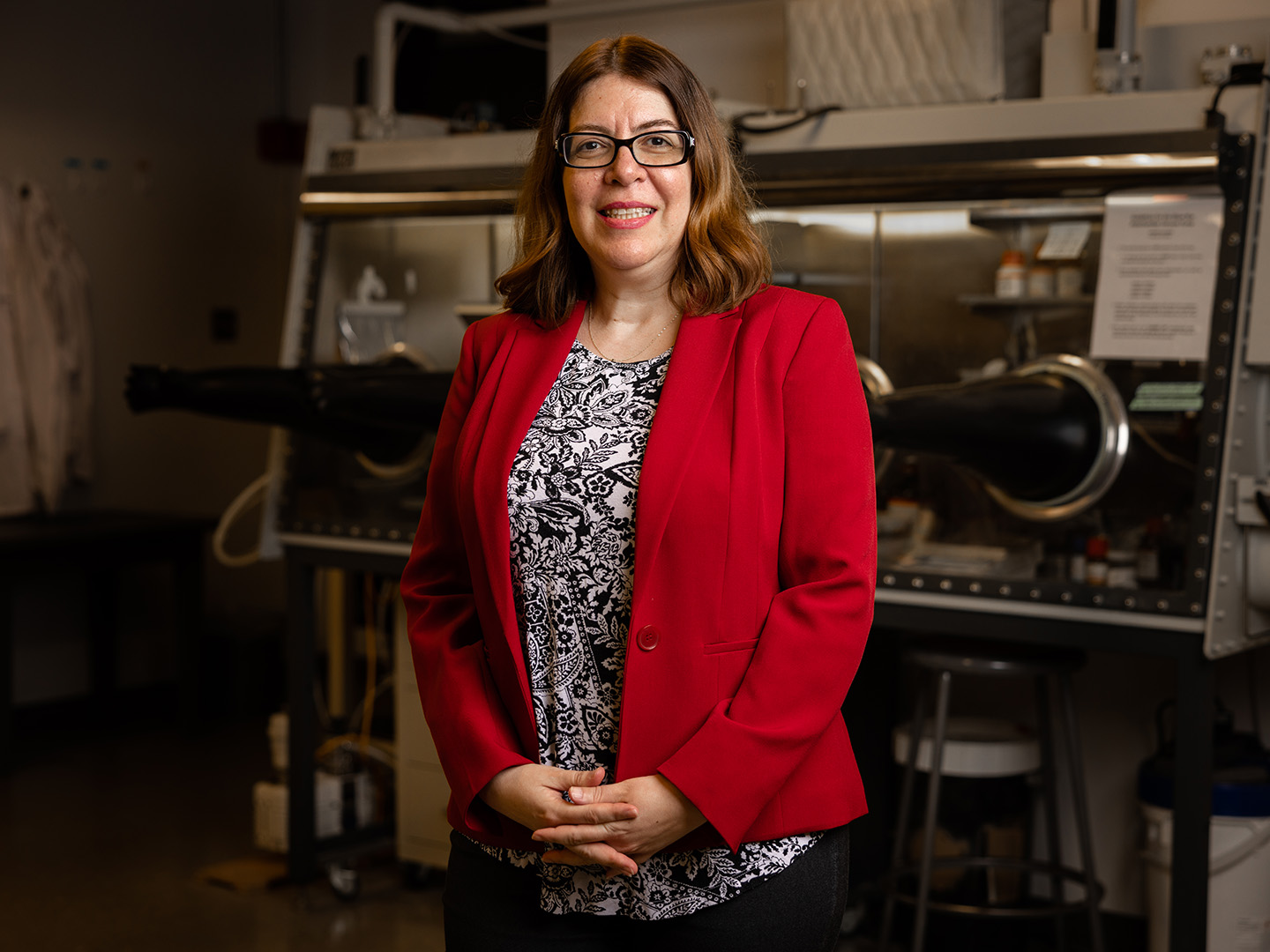 A woman in a red blazer stands in front of scientific equipment, smiling and posing for a photo.