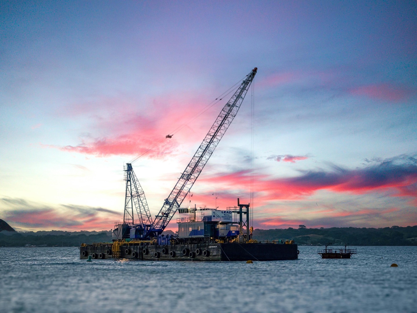 Crane on a barge in water under a pink and blue sky.