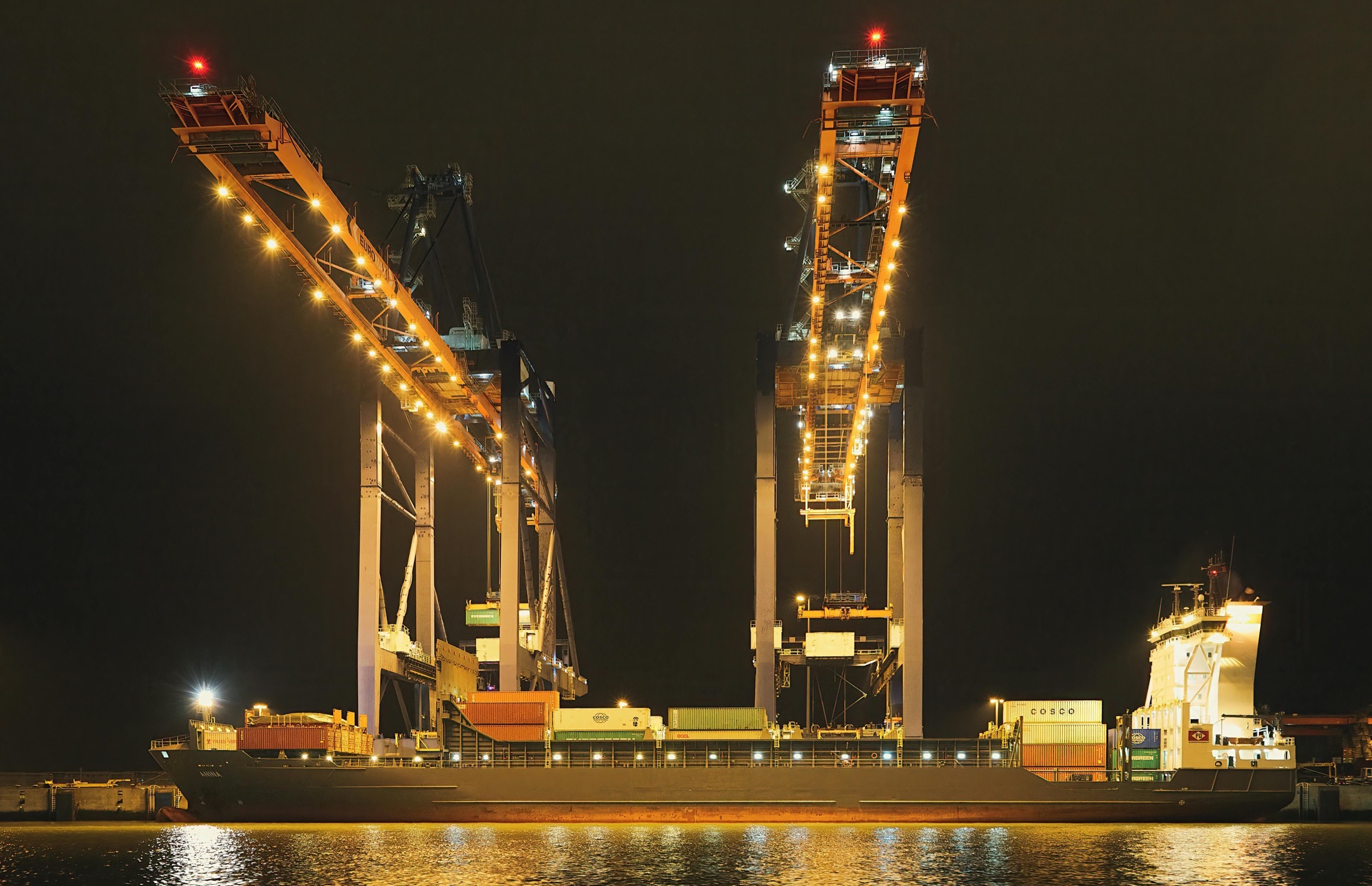 Cargo ship with stacked containers docked at night under bright lights.