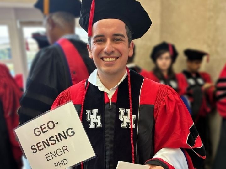 Graduate in red and black regalia smiles while holding a sign that reads “Geo-sensing engr PhD” at a University of Houston ceremony.