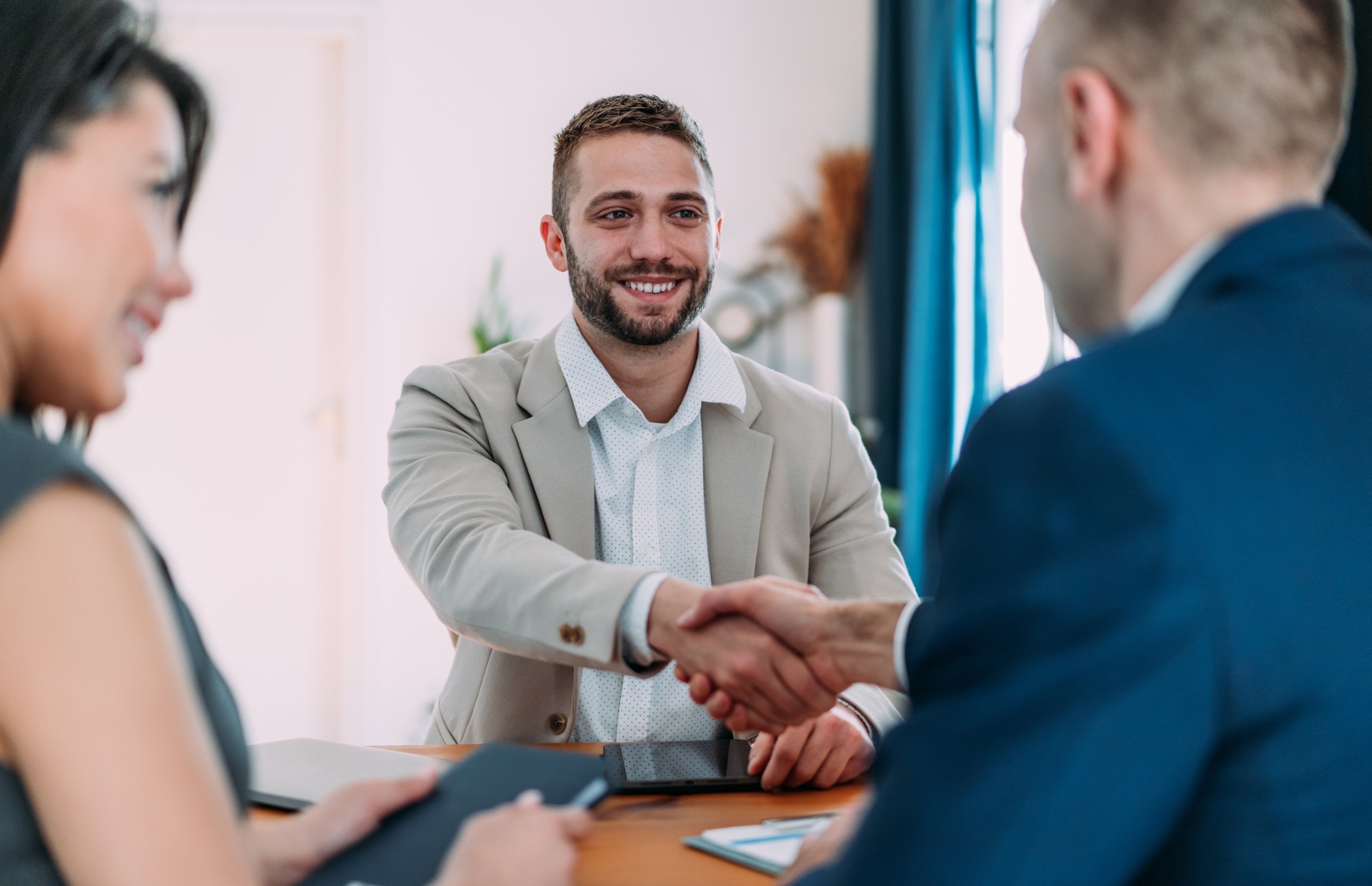 Man in beige suit shaking hands with another man in blue suit at a table.
