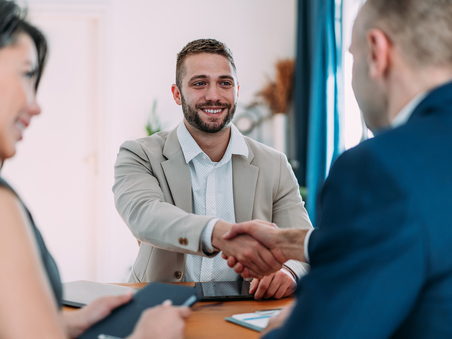 Man in beige suit shaking hands with another man in blue suit at a table.