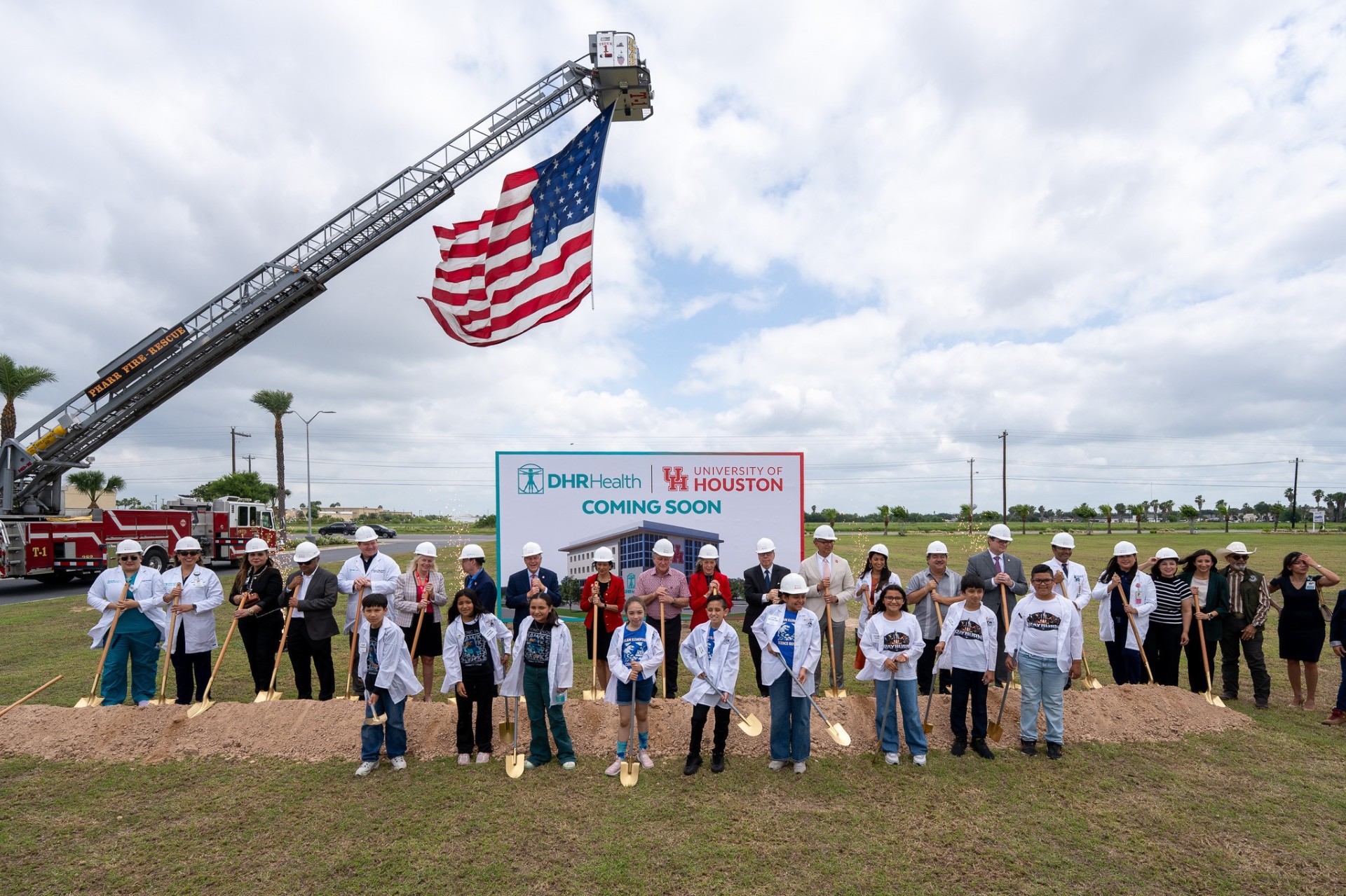 Groundbreaking ceremony with a group of people wearing hard hats and holding shovels. They stand in front of a DHR Health and University of Houston sign that says "Coming Soon." An American flag is displayed on a fire truck ladder in the background.