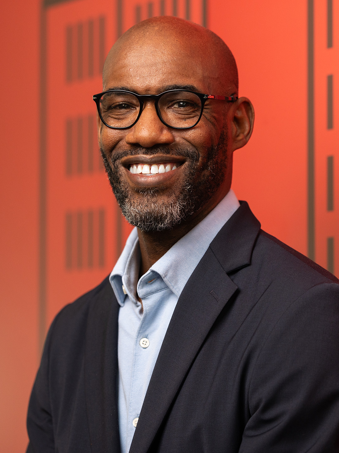 Bald man in glasses smiling against red background.