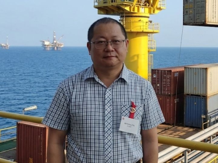 Weihang Zhu Man in glasses wearing name tag standing on ship deck with shipping containers and oil rigs.