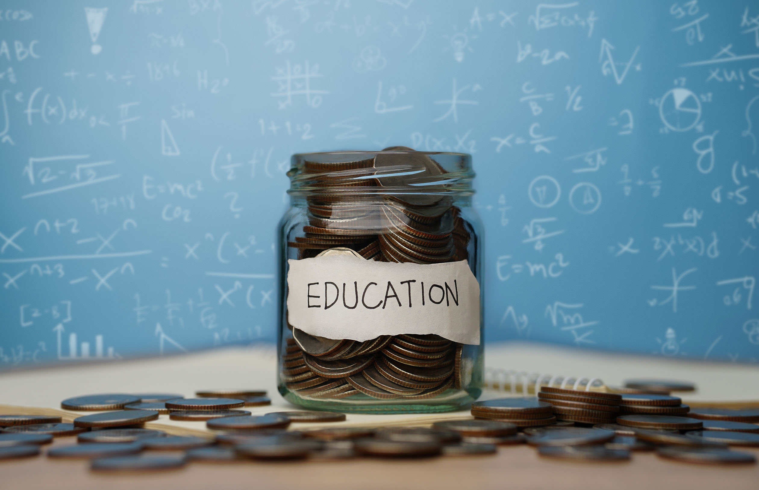 A glass jar labeled "Education" is filled with coins. More coins are scattered around the jar on a table. In the background, a blue board is covered with various mathematical equations.