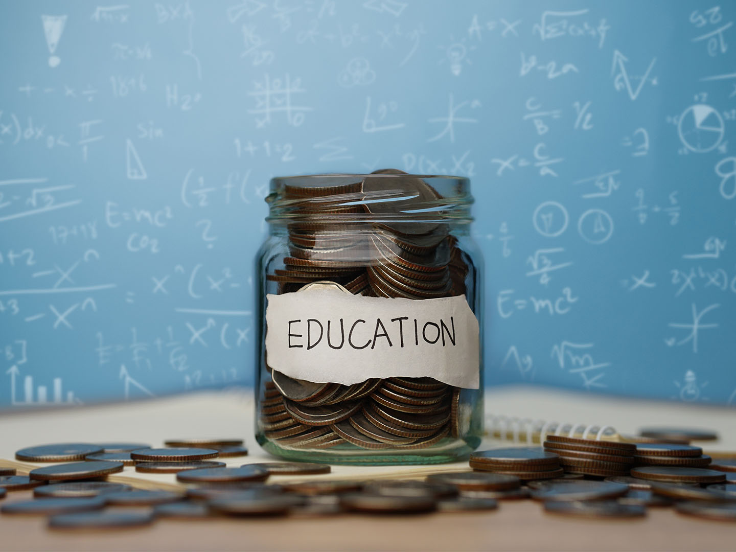 A glass jar labeled "Education" is filled with coins. More coins are scattered around the jar on a table. In the background, a blue board is covered with various mathematical equations.