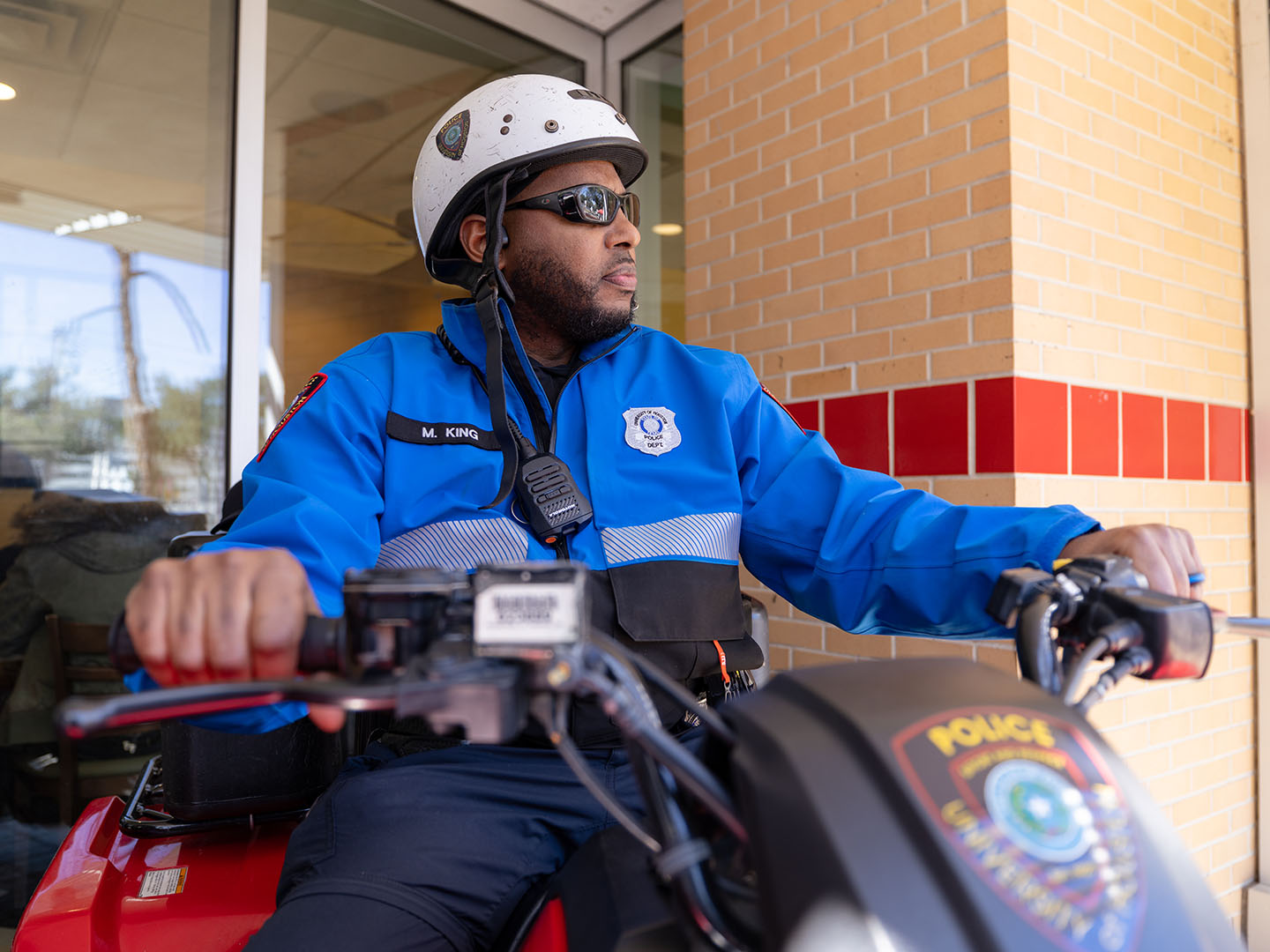 A person wearing a blue and black uniform with a helmet and sunglasses is sitting on a police vehicle. The uniform has a patch reading "Police" and a name tag.