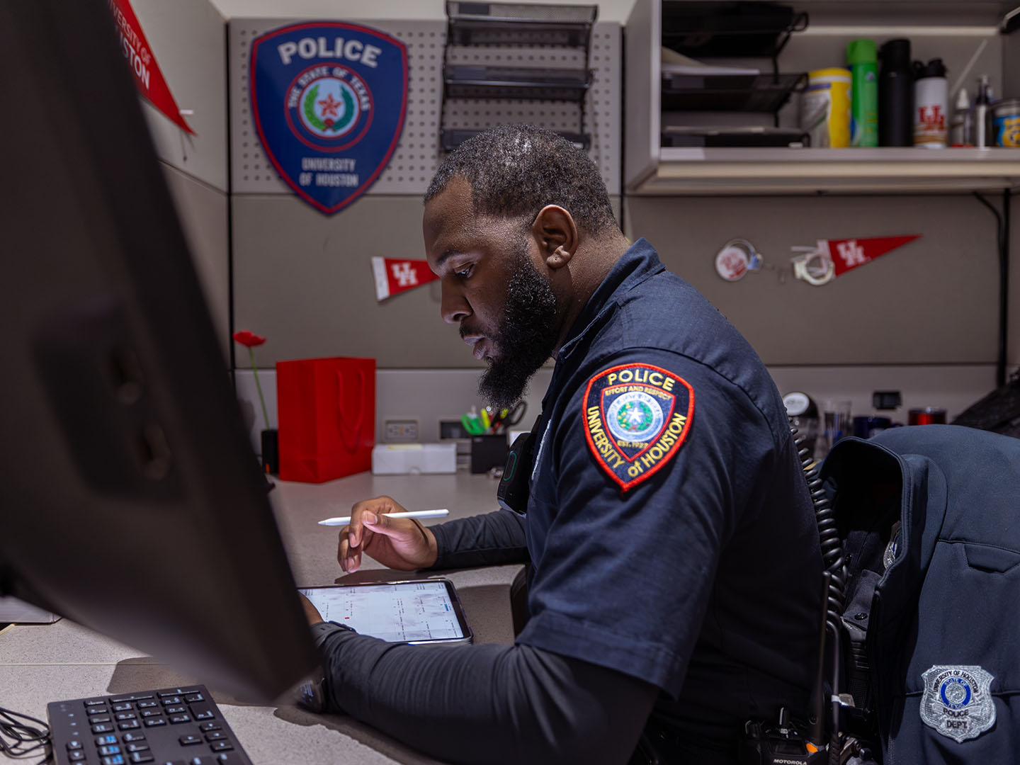 A person in a University of Houston police uniform is seated at a desk, working on a tablet. The office cubicle is decorated with University of Houston banners and a police badge.