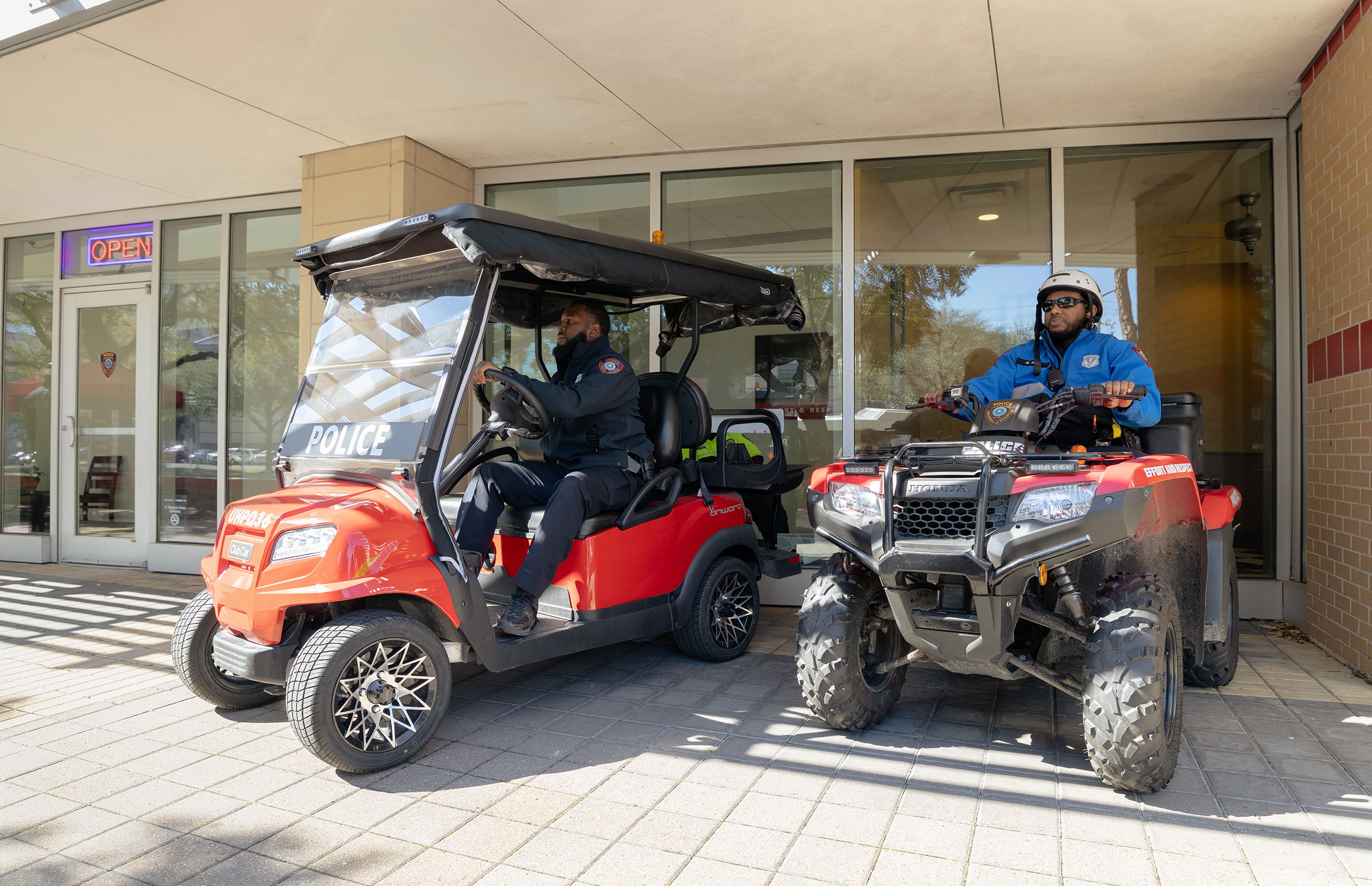 Two individuals in police uniforms are sitting on vehicles parked outside a building with large windows. One is on a red golf cart labeled "Police," and the other is on a red ATV. A sign with "Open" is visible on the window of the building.