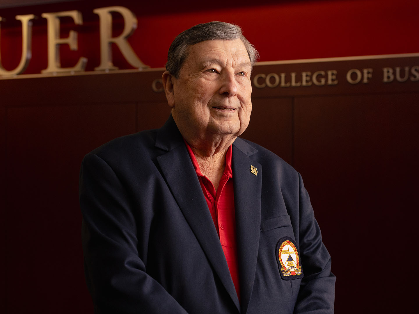 A person wearing a dark blazer with an embroidered badge stands in front of a wall with the words "Bauer College of Business." The person is smiling slightly.