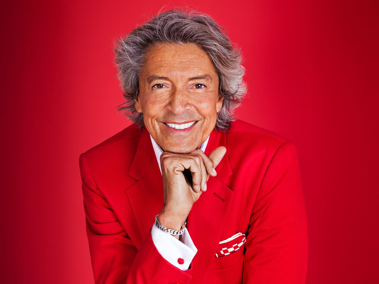 Tommy Tune wearing a red suit while resting smiling face above hand in a posed portrait