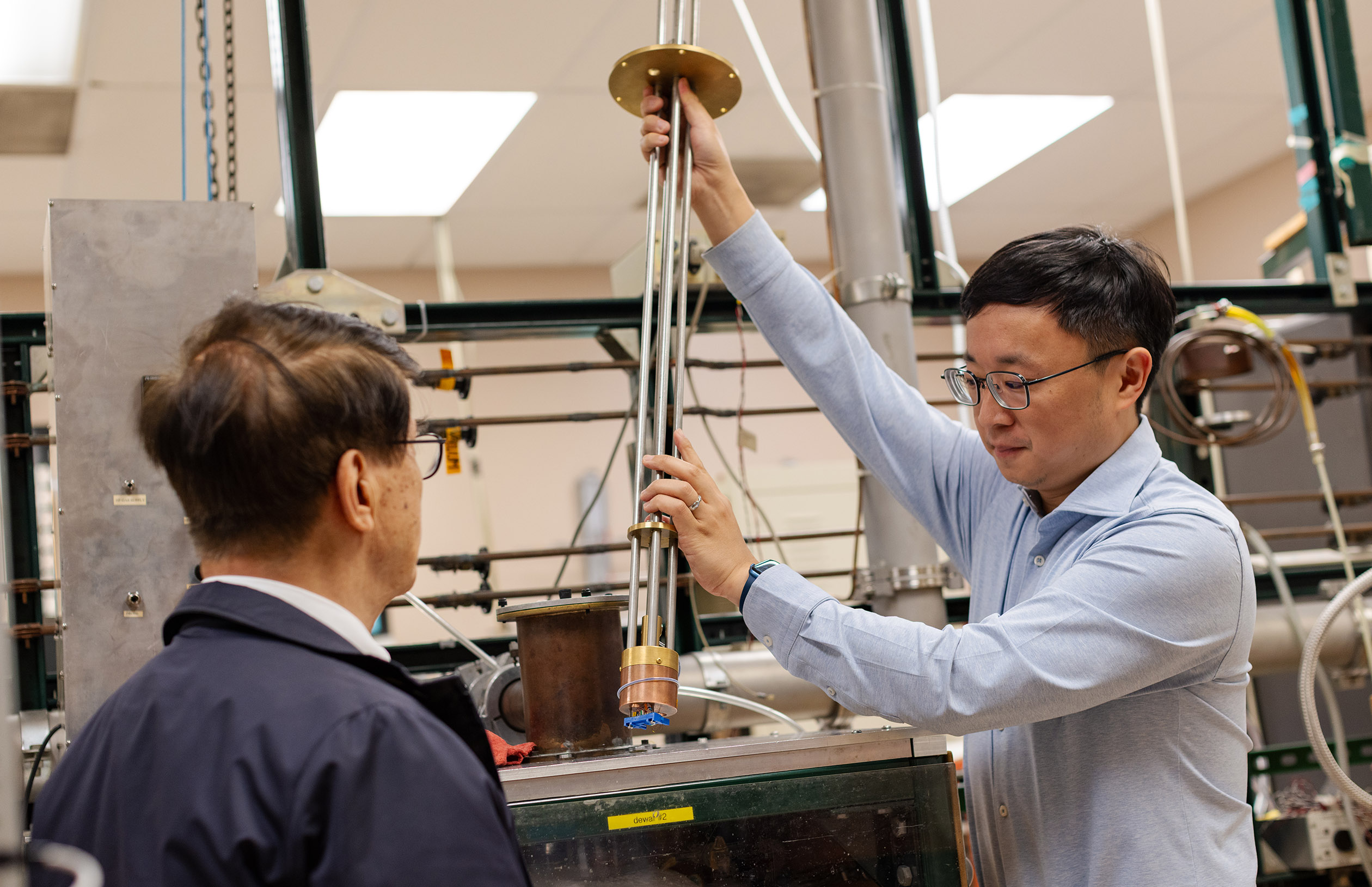 Two individuals are in a laboratory setting. One is holding a long metal rod connected to equipment, while the other observes. The space is filled with various scientific apparatus and machinery.