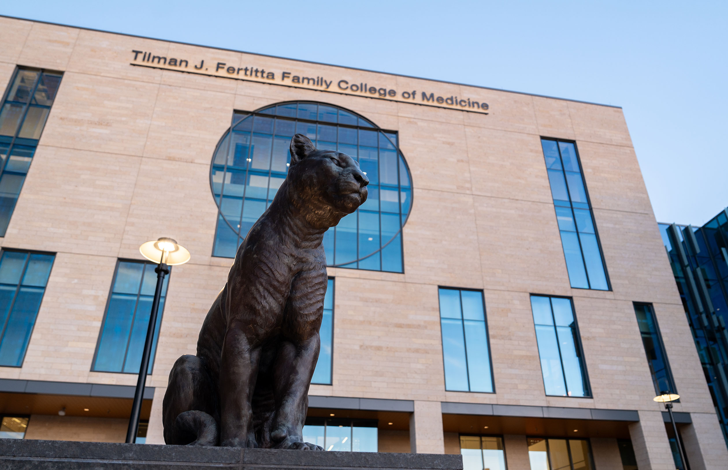 Bronze cougar statue in front of Fertitta College of Medicine building with a circular window and text above.