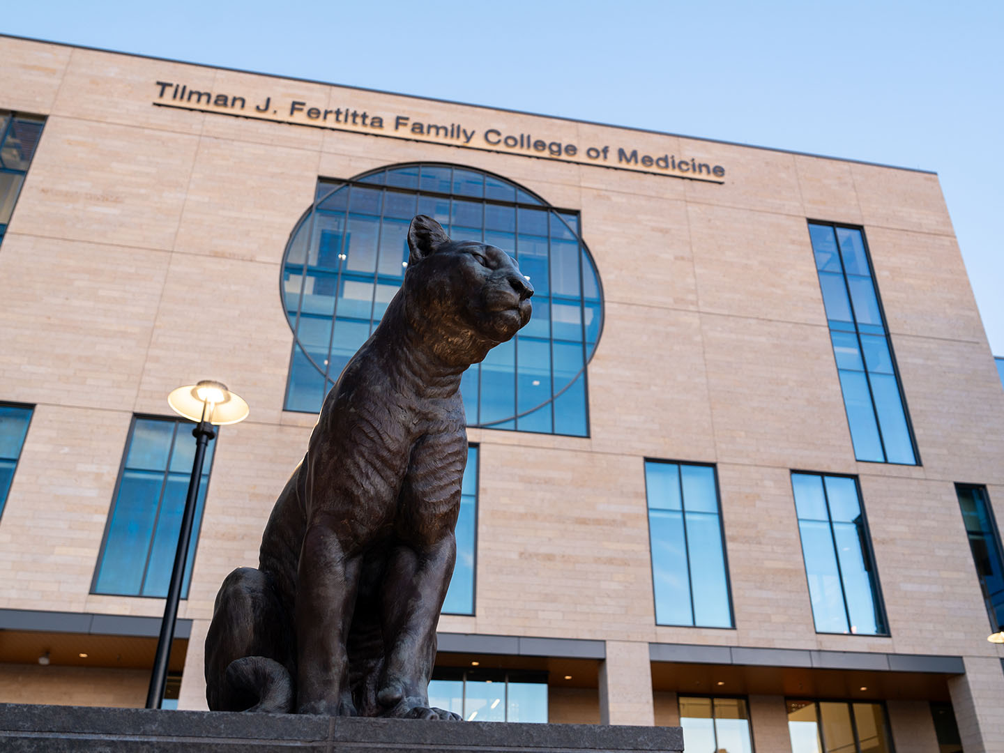 Bronze cougar statue in front of Fertitta College of Medicine building with a circular window and text above.