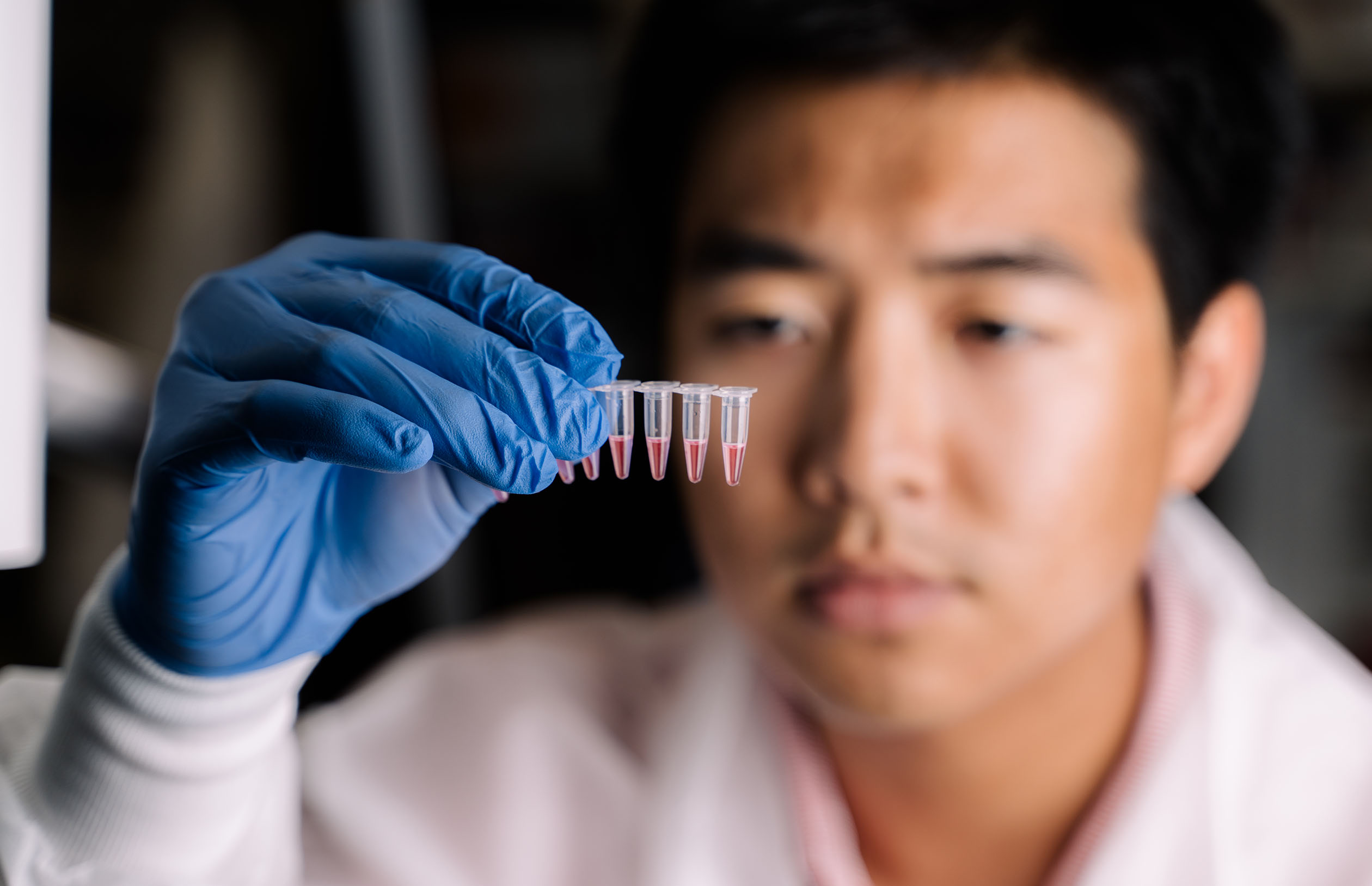 Brandon Than in a lab coat and blue gloves holding a test tube rack with pink liquid.