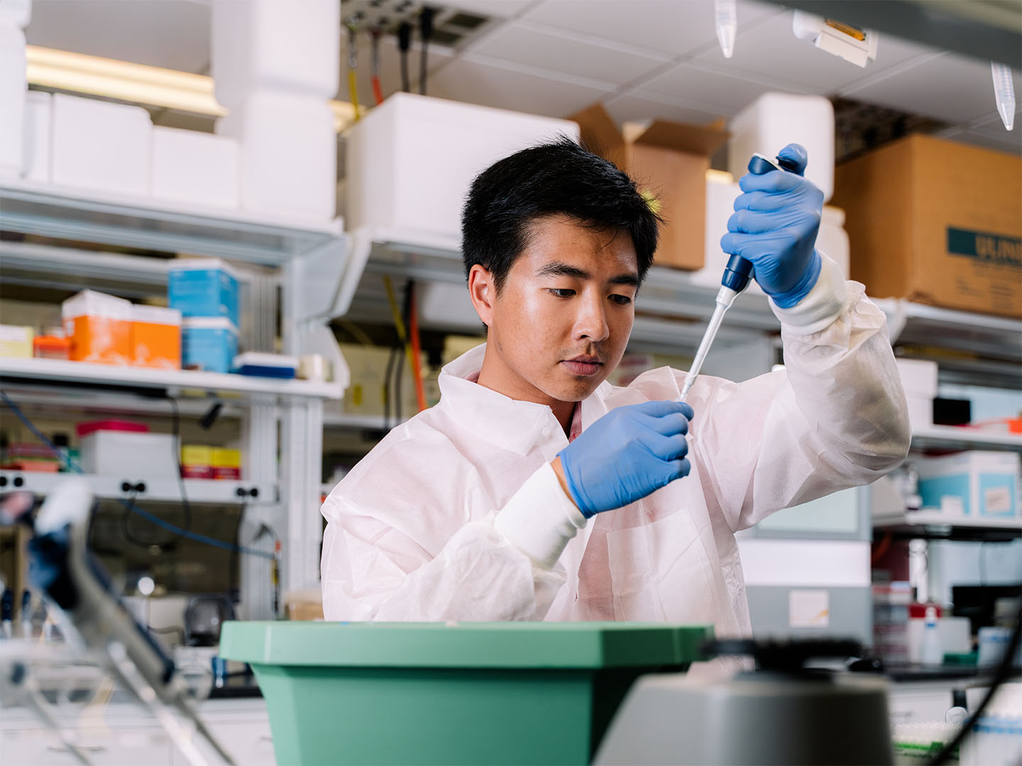 Brandon Than in a lab coat and blue gloves using a pipette in a laboratory.