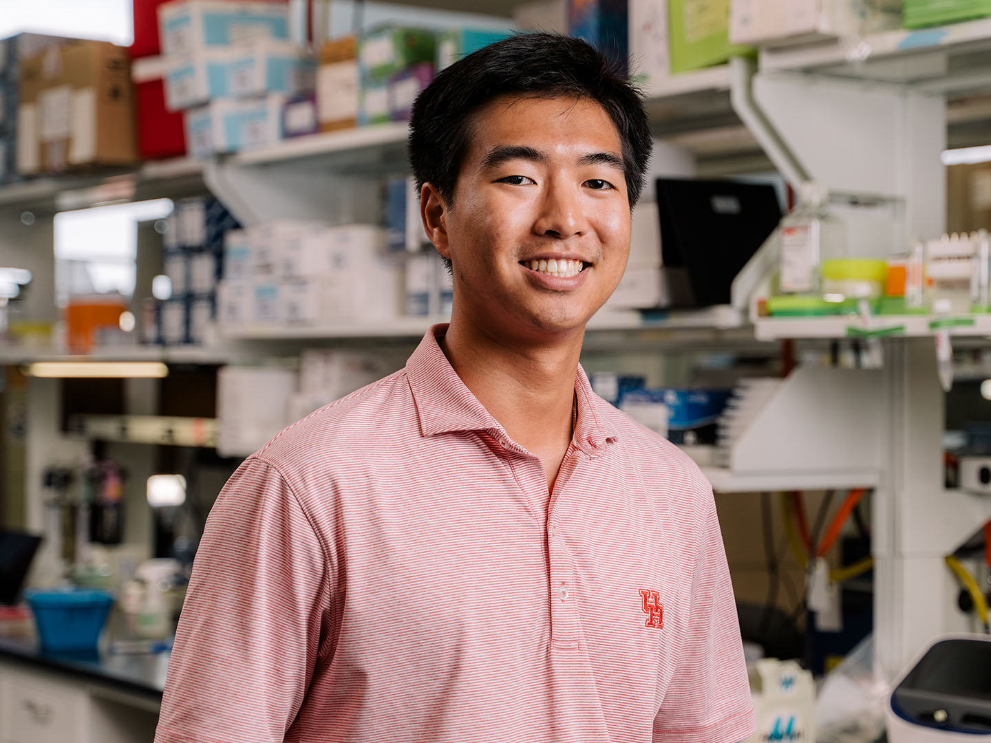 Brandon Than in red polo shirt smiling in laboratory with shelves of boxes and equipment.