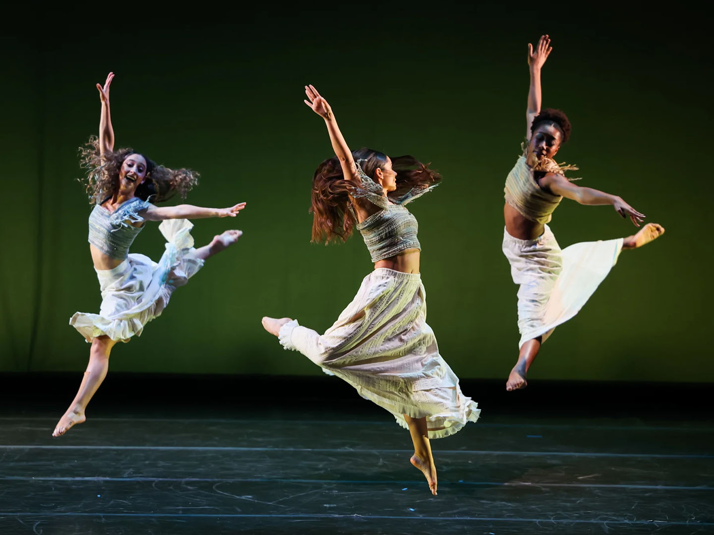 Three women in white dresses leaping in the air on a stage.