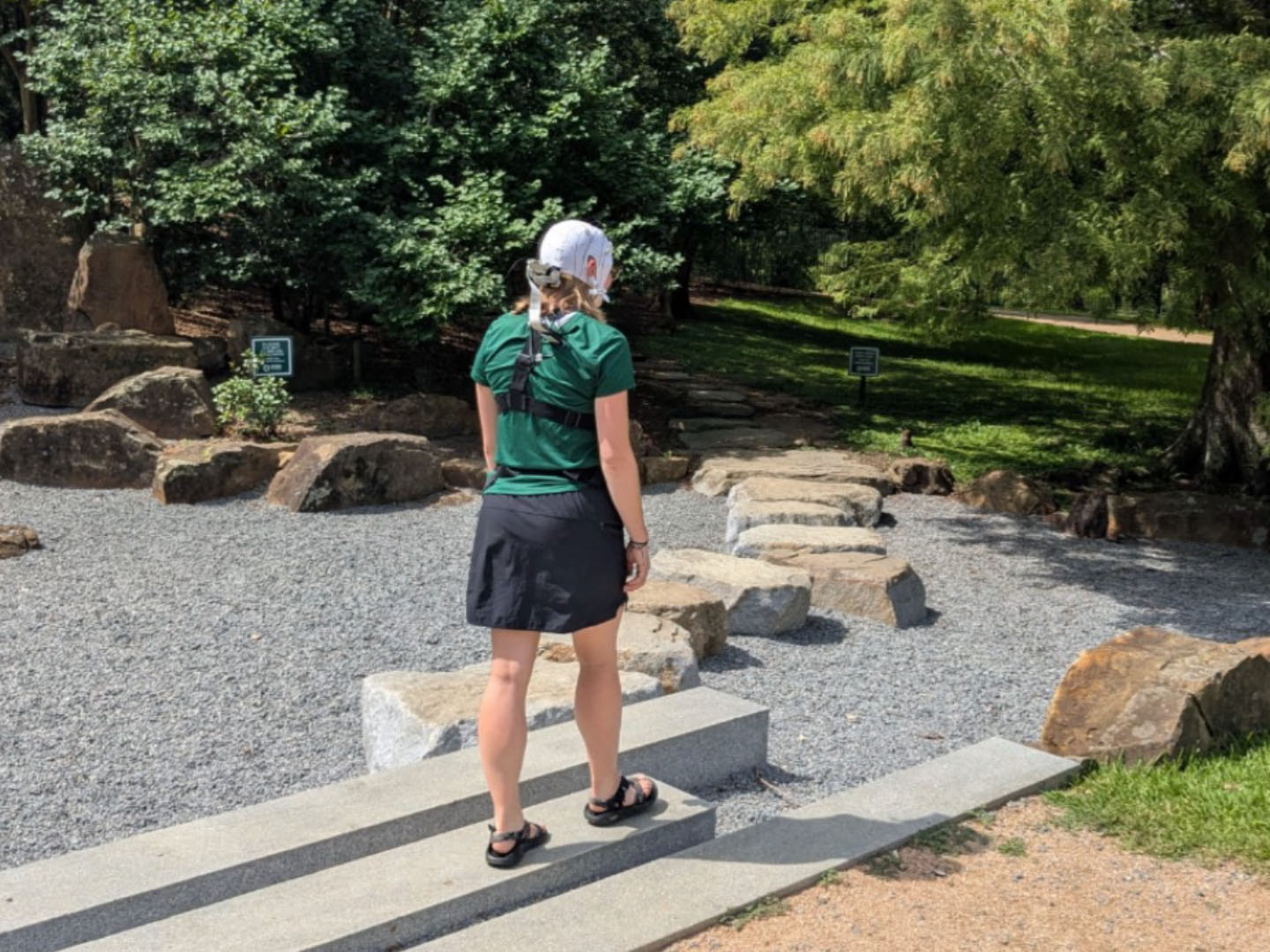 A woman in a brain cap stands on concrete steps, facing a stone path in a wooded area.
