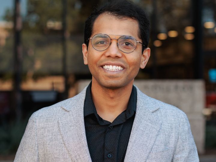 Man wearing glasses and a gray blazer smiles outdoors in front of a blurred building entrance.