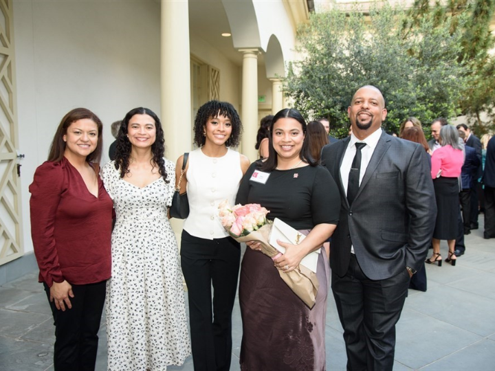 Five adults pose together outdoors at a formal event, with one woman holding a bouquet of pink roses and a folder.