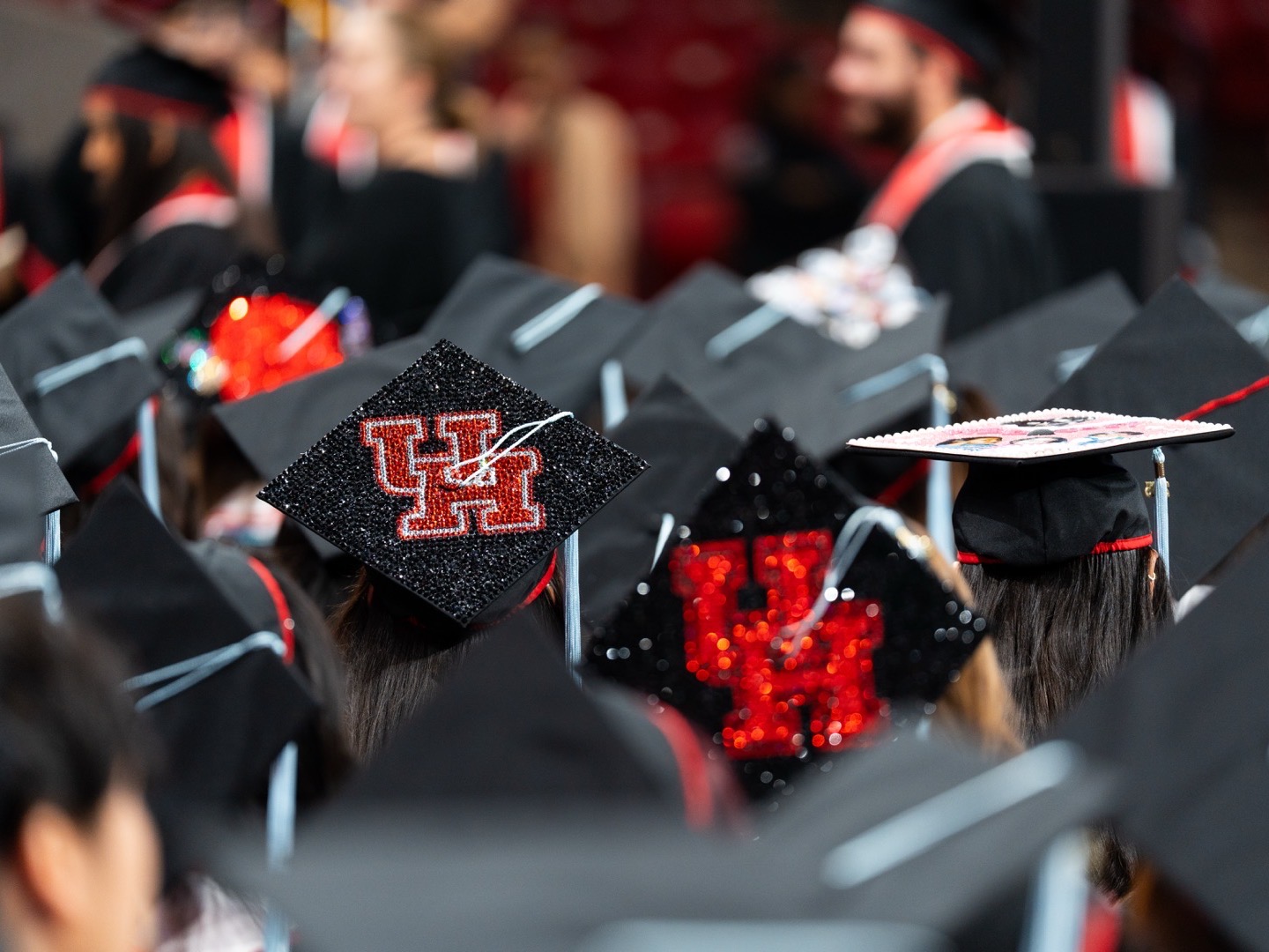 Graduates in black caps sit in rows, with several caps decorated in black and red glitter showing the letters UH.