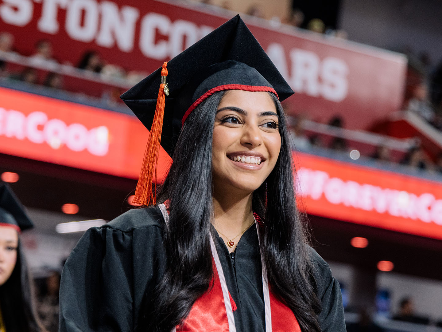 Graduate in a black cap and gown smiles during a ceremony with a red “Houston Cougars” sign in the background.