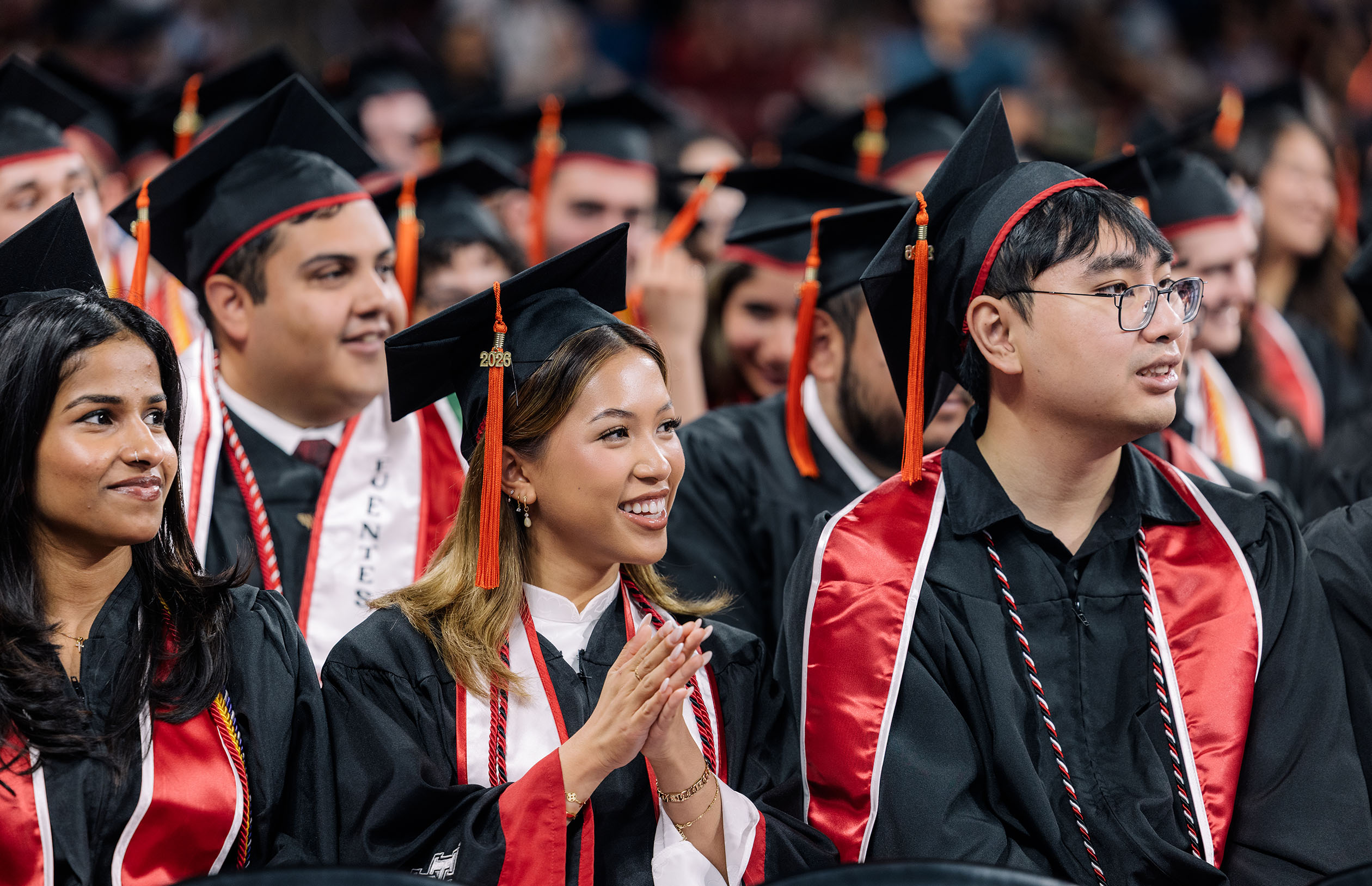 Graduates in black gowns and caps with orange tassels smile and clap during the 2026 commencement ceremony.