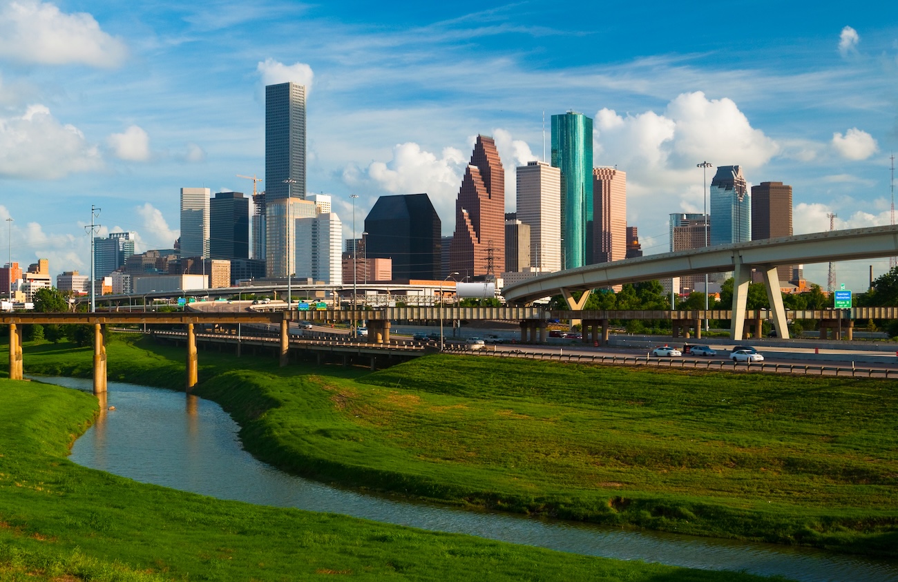 Houston skyline rises behind highways and a green waterway under a bright blue sky with scattered clouds.