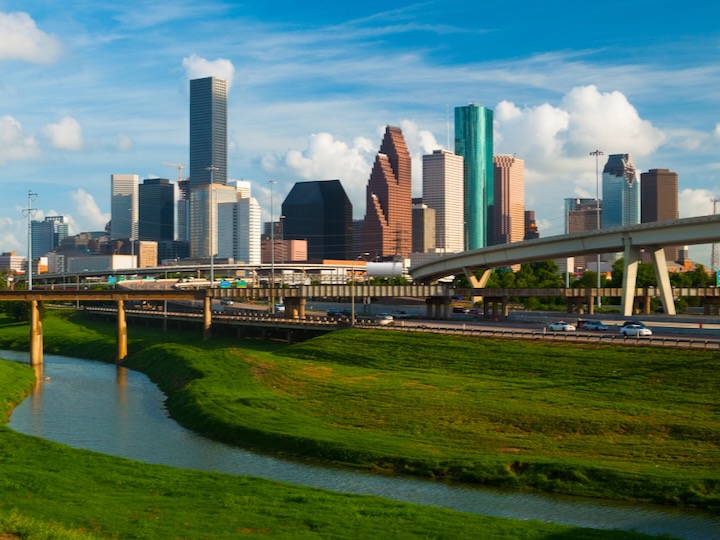 Houston skyline rises behind highways and a green waterway under a bright blue sky with scattered clouds.