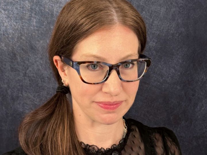 Woman with brown hair in a low side ponytail wears tortoiseshell glasses and a black lace top against a dark backdrop.