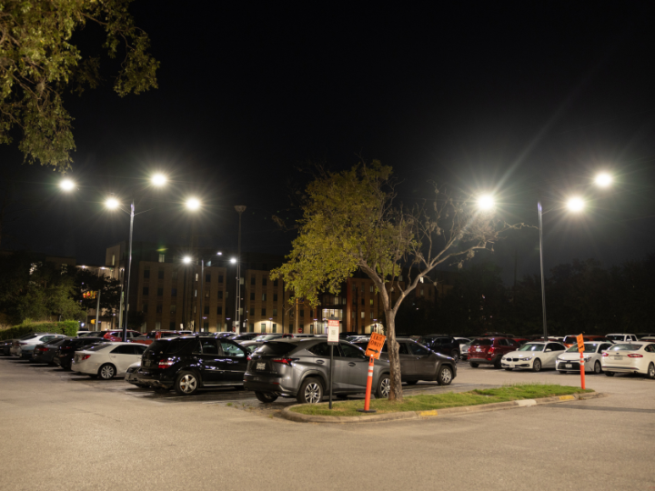 A well-lit parking lot at night shows rows of parked cars under bright streetlights.