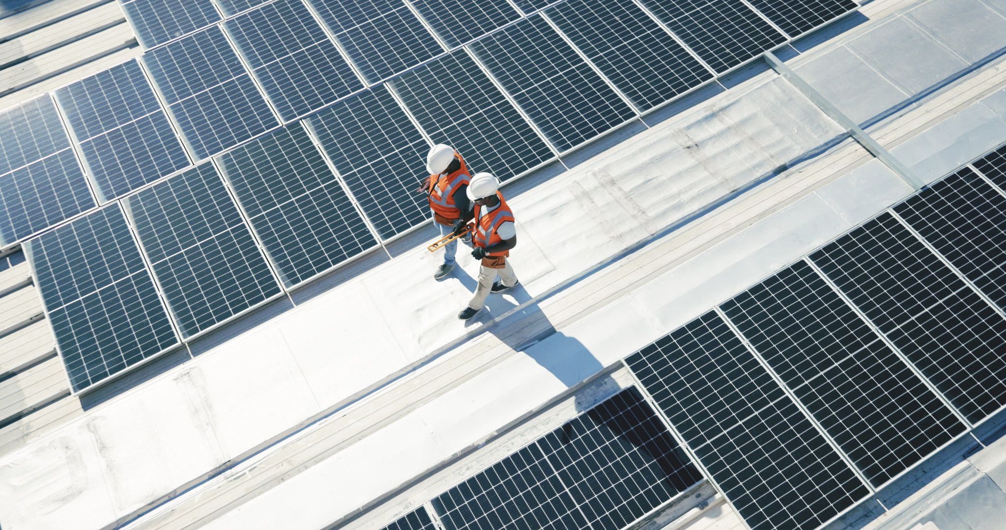Two workers in safety gear inspect rooftop solar panels under bright sunlight.