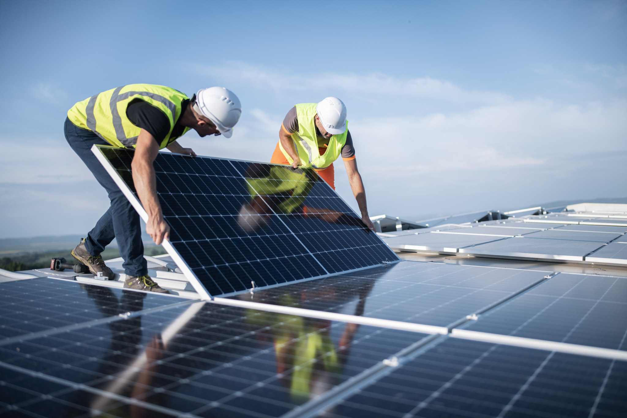Workers in safety vests and helmets install solar panels on a rooftop under a clear blue sky.