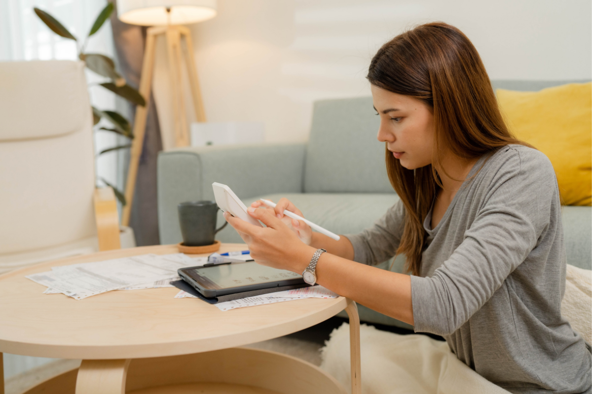 Woman calculates expenses at a low table with bills, a tablet, and a calculator in a living room.