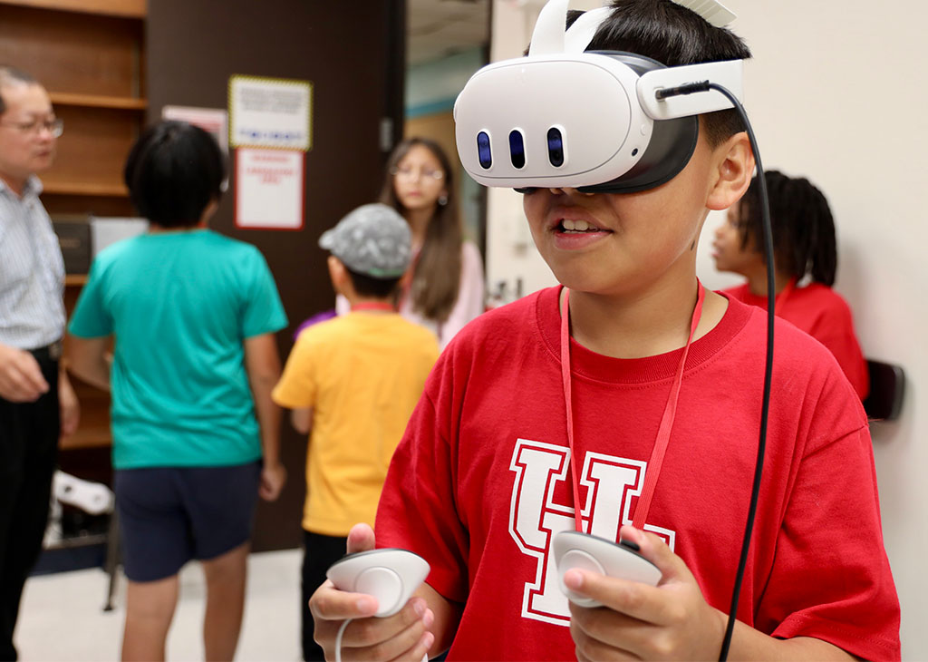 A student wearing a University of Houston T-shirt and a virtual reality headset uses handheld controllers during a classroom activity.
