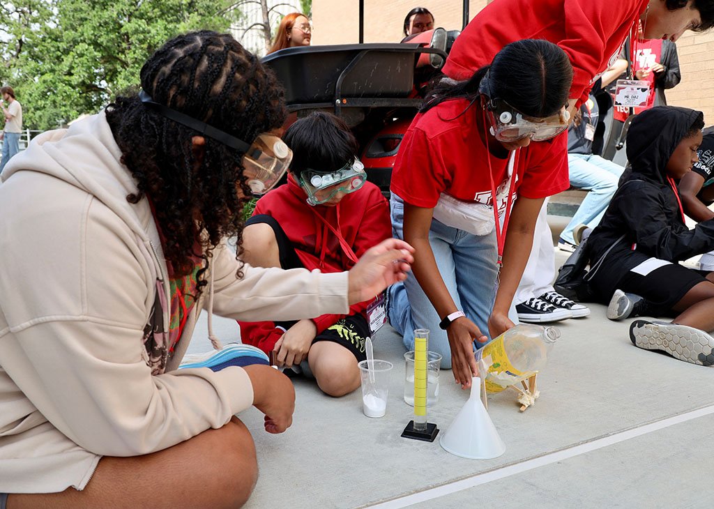 Students wearing safety goggles conduct an outdoor science experiment, pouring liquid into a funnel and graduated cylinder while recording observations on paper.