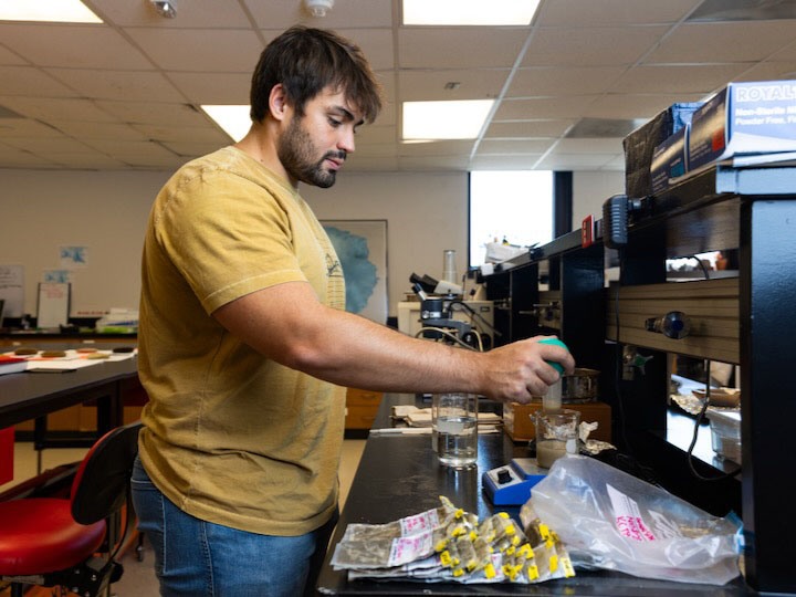 Graduate student Breno Araujo uses a large pipette to gather sediment samples mixed by a magnetic stirrer.