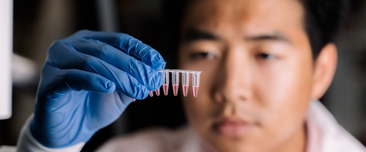 Brandon Than in a lab coat and blue gloves holding a test tube rack with pink liquid.