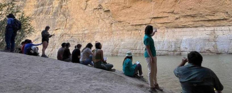 Students gathered on the banks of a river looking at a rock outcrop.