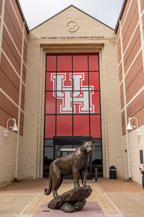 A bronze cougar statue stands in front of a building entrance. Above the entrance is a large red banner with the interlocking UH logo.