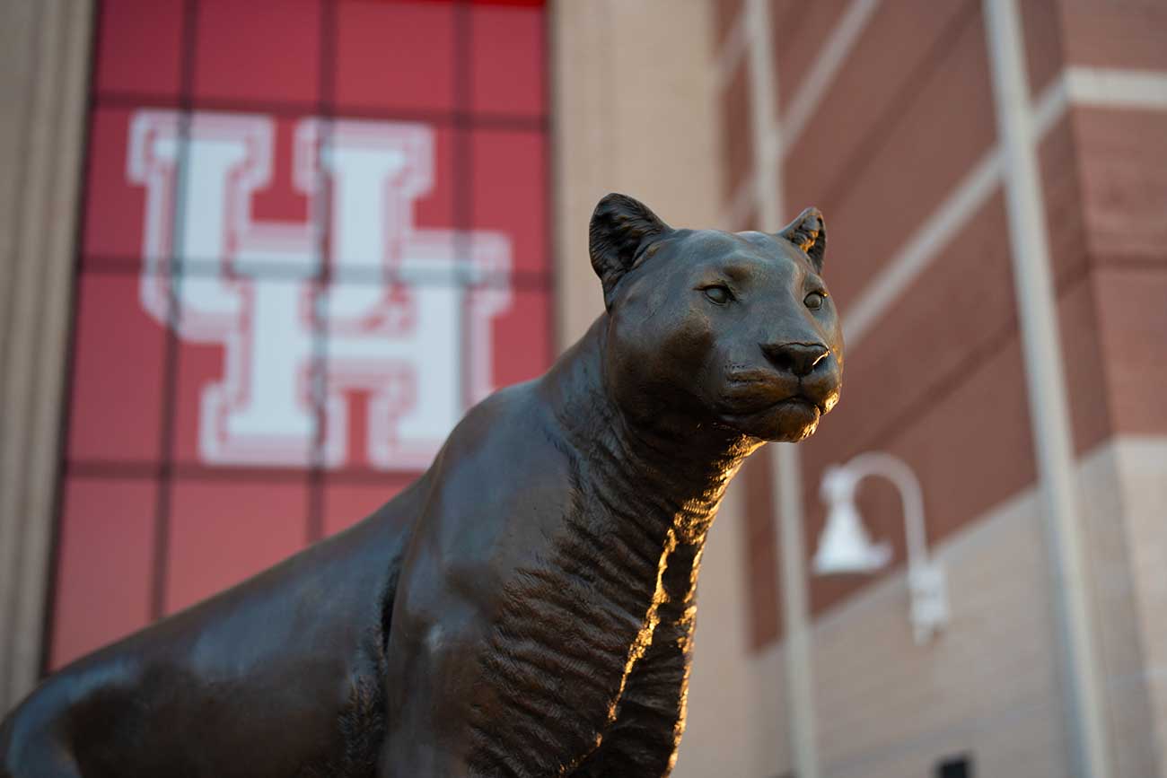 Photograph of a bronze cougar statue facing right. A large red banner against a building is in blurred in the background.