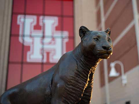 Photograph of a bronze cougar statue facing right. A large red banner against a building is in blurred in the background.
