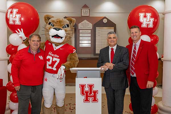 Photograph of three men and a cougar mascot standing by a white lectern flanked by ballon decorations.