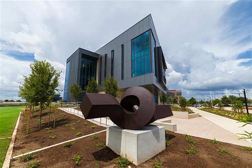 A metalic sculpture of a brown elongated rectangular prism twisted in a loop rests on platform at the corner of a grey building.