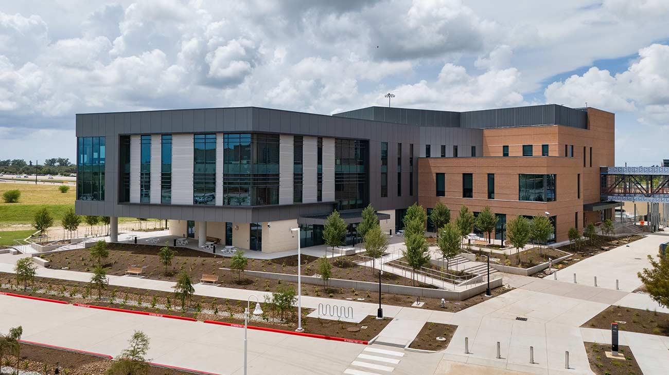Photo of the exterior of a three story building with brown bricks and dark grey sides. Newly planted trees fill the soil surrounding the walkways.