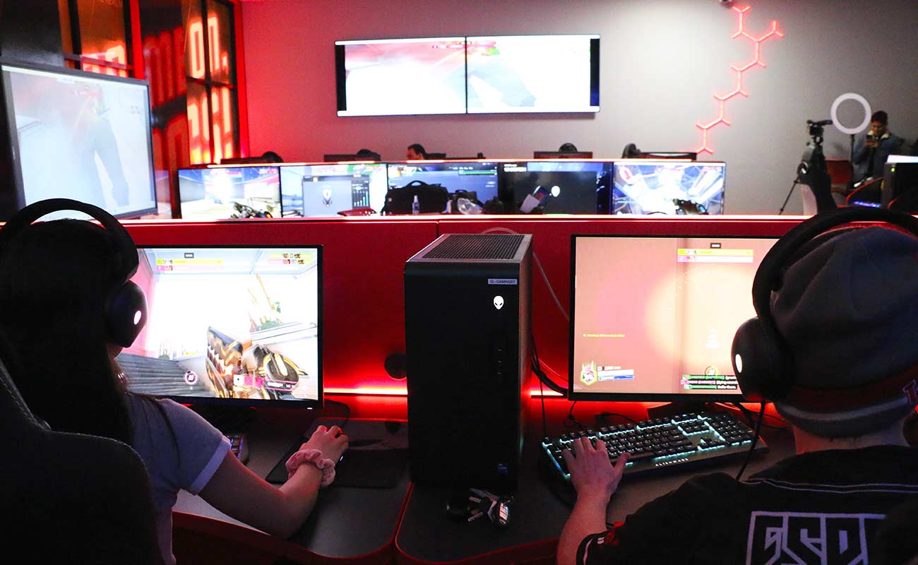 A team of gamers sit in front of computers in a darkened room with multiple monitors on the wall.