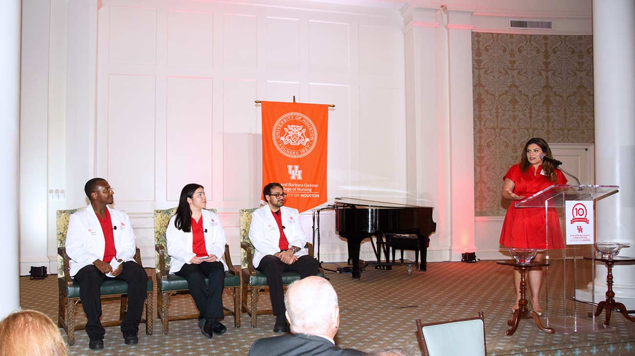Nursing students in lab coats sit together in a ballroom next to a woman in a red dress speaking at a podium.