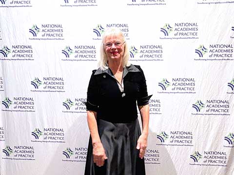 A blonde woman in a black evening gown and jacket smiles in front of a white backdrop with the National Academies of Practice logo.